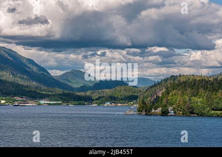 Ketchikan, Alaska, USA - July 17, 2011: Wide landscape of surrounding forested mountains under heavy cloudscape. Buildings on shoreline add color. Blu Stock Photo