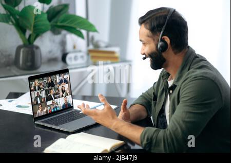 Successful busy mixed race successful man, sitting at work desk in modern office, using laptop talking to multiracial business team via video conference. Virtual online meeting, remote negotiations Stock Photo