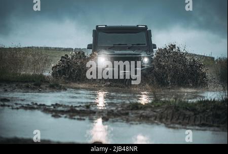 Water splash in off-road racing. Classic 4x4 car crossing water with splashes on muddy road. Low angle view of front of SUV on mountain road. Stock Photo