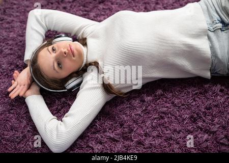 A young girl is thoughtfully listening to music while lying on a purple carpet. Stock Photo