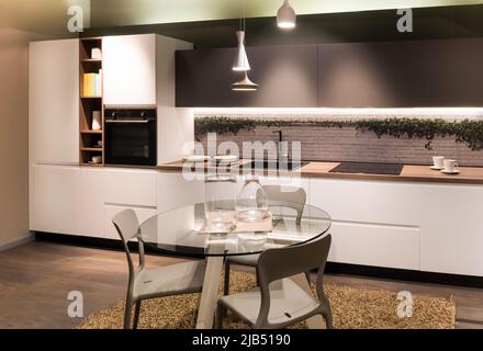 Round glass table with glassware and chairs located near cupboards and counters with built in appliances in modern kitchen at home Stock Photo