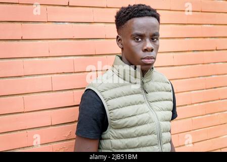 Serious African man in vest looking at camera while standing near orange brick wall on city street Stock Photo
