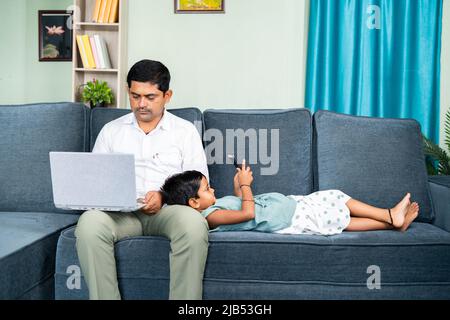 Father working on laptop while kid using mobile phone by sleeping on father lap at home - concept of togetherness, internet and technology lifestyle Stock Photo