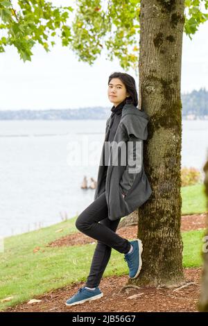 Smiling biracial teen girl or young adult female in gray jacket leaning against tree in park by lake Stock Photo