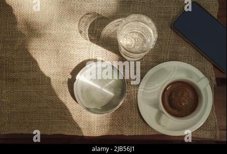 Morning cup of coffee, glass of water, mobile phone and ash tray on old wooden table, looked from above. Stock Photo