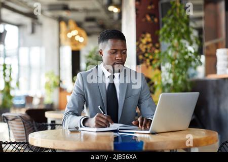 Serious busy young African-American entrepreneur in gray suit sitting at table and planning tasks, he making notes in diary while using laptop in cafe Stock Photo