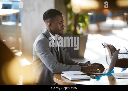 Serious busy young African-American businessman with beard sitting at table and typing on laptop while browsing internet Stock Photo