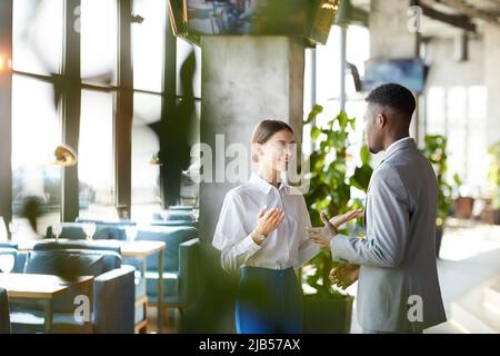 Positive confident multi-ethnic business colleagues in formalwear standing in modern restaurant and gesturing hands while discussing strategy Stock Photo