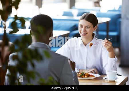 Positive excited young business lady in white shirt sitting at table and eating pasta while meeting with black executive in cafe Stock Photo