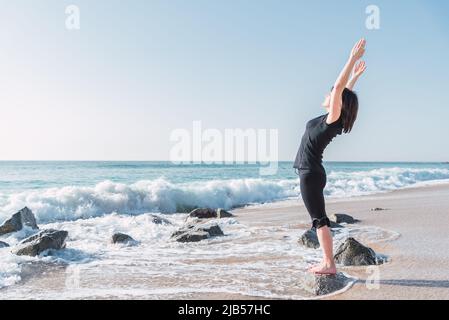 Side view of female stretching body and doing backbend while standing on rock in Tadasana. She is with raised arms on sea coast against blue sky during yoga session Stock Photo