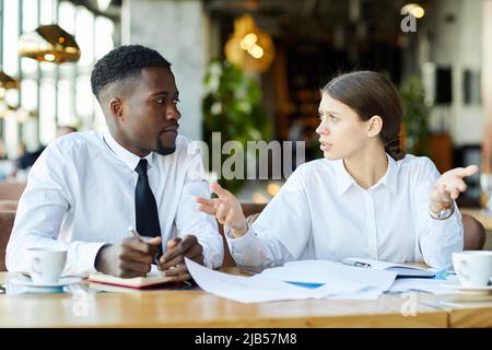 Puzzled young multi-ethnic business colleagues in white shirts sitting at table and discussing report data while working with papers in restaurant Stock Photo
