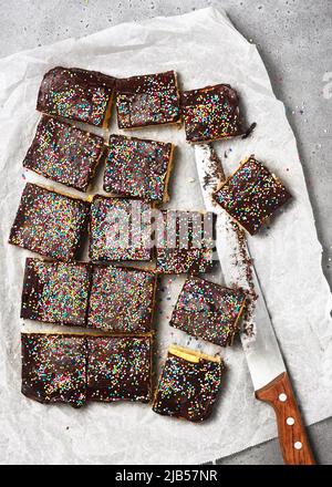 Delicious biscuit cake with dark chocolate icing and colorful sugar sprinkle in bars on baking paper with knife. Homemade sweet food concept. Top view Stock Photo