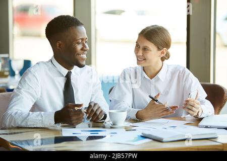Positive young multi-ethnic business analysts sitting at table with papers and working with financial data while discussing development strategy Stock Photo