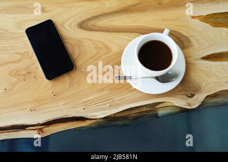 High angle view of black coffee in cup with spoon and smartphone placed on table with wooden structure Stock Photo