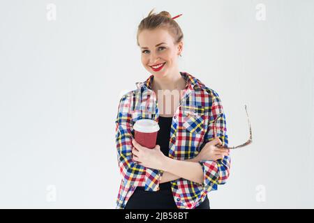Thoughtful young woman wearing checkered shirt standing with her hands folded across the chest, holding coffee and sunglasses Stock Photo