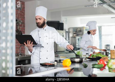 Gastronomic expert preparing dish garnish using fresh organic ingredients while holding laptop with one hand. Gourmet cuisine chef cooking dinner dish while using computer to follow recipe closely Stock Photo