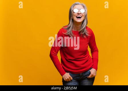 Charming senior woman in sunglasses laughing toothy. Elegant carefree mature modern gray-haired lady making looking at sun, enjoying sunlight isolated over yellow background Stock Photo