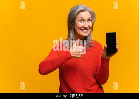 Delighted by the holiday deal mature woman holding smartphone with empty screen. Overjoyed middle-aged lady demonstrates phone with blank display and showing big finger isolated on yellow Stock Photo