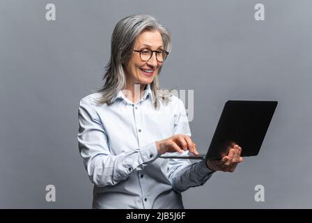 Confident modern elderly woman using laptop isolated on grey. Retirement female entrepreneur typing, middle-aged lady with gray hair in casual wear checking emails Stock Photo
