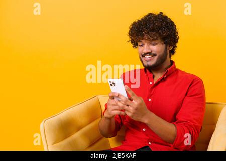 Waist up view of happy handsome blogger young adult man sitting at the armchair, using smartphone and texting message with smile. Indoor, isolated, studio shot, orange background Stock Photo