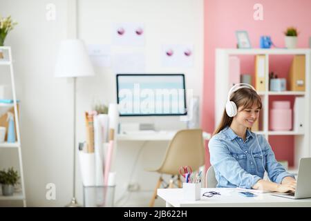 Cheerful young lady in wired headphones sitting at table and using laptop while working with pleasure on web project Stock Photo