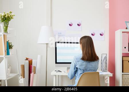 Rear view of brunette lady in denim shirt sitting in front of modern computer and preparing presentation on business analysis Stock Photo