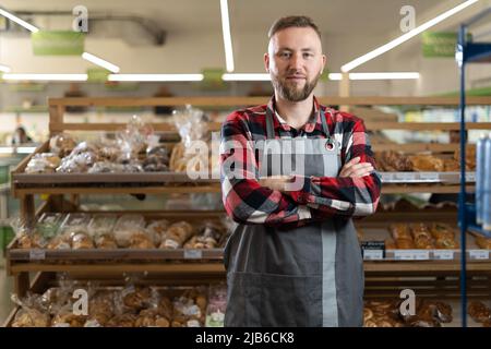 Serious baker standing with fresh bread in a bakery, caucasian man standing with crossed arms at the bakery counter and looking at the camera, tired Stock Photo