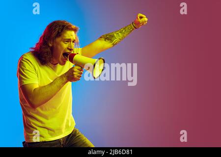 Young excited man with long curly hair shouting at megaphone isolated on gradient blue-pink background. Concept of beauty, fashion, youth culture and Stock Photo