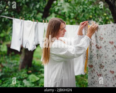 middle aged caucasian woman in white dress doing homework, hanging clothes on clothesline on street in courtyard of village cottage house, laundry day concept Stock Photo