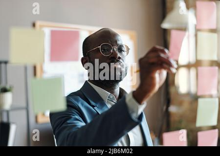 Serious African businessman in suit and eyeglasses making notes on sticky notes hanging on glass wall at office Stock Photo