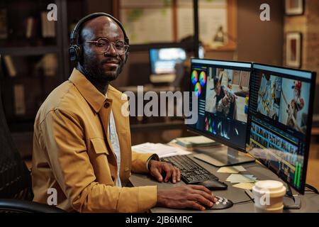 Portrait of African american colorist in headphones looking at camera while sitting at table with pc and making visual content Stock Photo