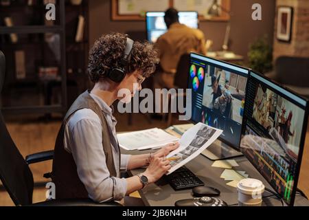 Female editor in headphones examining script at her workplace while editing photos on computer Stock Photo