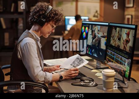 Young footage editor in headphones examining script during editing of video on computer with editing software Stock Photo