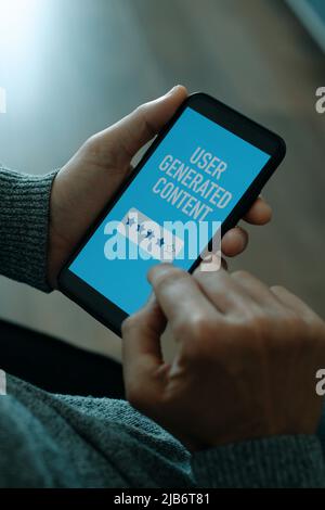 closeup a young man sitting in a chair holding his smartphone, that reads the text user generated content in its screen, in his hand Stock Photo
