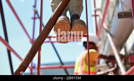 Two construction workers on scaffolding platform core drilling in sunshine. Steel toe capped work boot soles on red metal ladder rungs. Stock Photo
