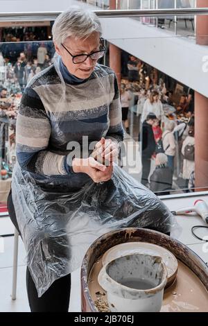 Senior gray-haired woman kneading clay in her hands to sculpt clay vase on potter's wheel. Stock Photo