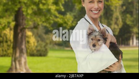 Image of happy caucasian female vet holding yorkshire terrier pet dog, over trees in park Stock Photo