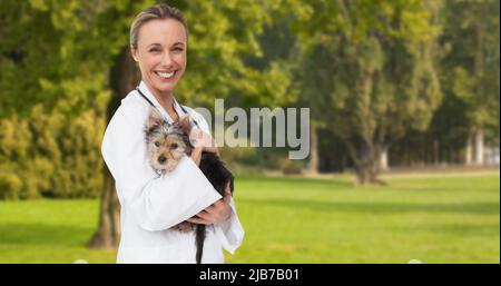 Image of happy caucasian female vet holding yorkshire terrier pet dog, over trees in park Stock Photo