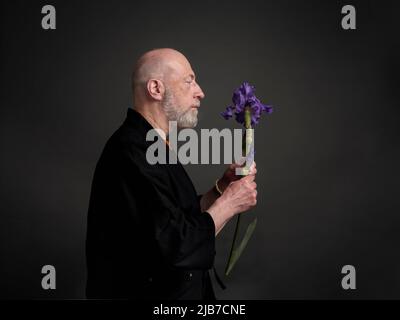 Head and shoulders profile portrait of bald and bearded senior man in kimono admiring an iris flower Stock Photo