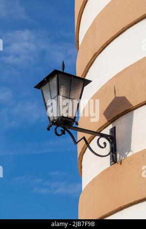 old, ornate iron street lantern hangs hangs on a round tower with sandstone and plaster facade in rings before strong blue sky Stock Photo