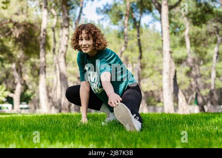 Beautiful young redhead woman wearing sportive clothes standing on city park, outdoors stretching leg muscles, doing side lunges in skandasana pose. H Stock Photo