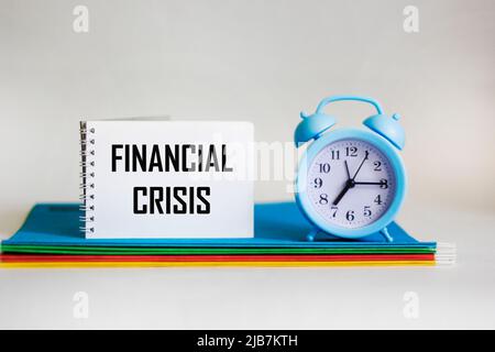 The word financial crisis is written on a notepad, which lies on multi-colored folders and next to a clock on a white background. Stock Photo
