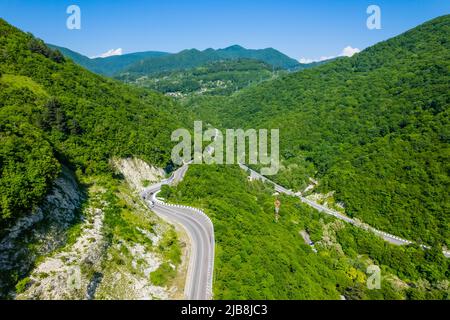 Winding road is meandering between blue sea and green mountains. Aerial view of car driving along the winding mountain road in Sochi, Russia. Stock Photo