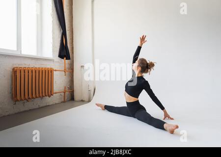 A young woman in black sportswear, stretches the muscles of the legs, performs a transverse split, sitting on a white cyclorama in the room Stock Photo