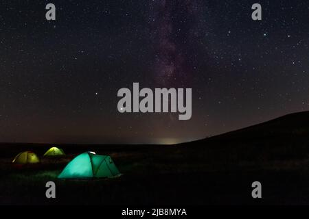 Travel and camping concept - three glowing camping tents at night under a starry sky with milky way and setting sun. Illuminated blue camping tent und Stock Photo