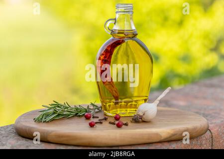 A glass cruet containing extra virgin olive oil flavored with red chili pepper is on a wooden cutting board, with garlic and rosemary Stock Photo