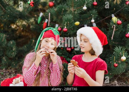 Merry Christmas. Portrait of two funny children girls in Santa hat eating gingerbread cookies drinking hot chocolate outside having fun. Happy Holidays. kids enjoying holiday. Christmas in July Stock Photo