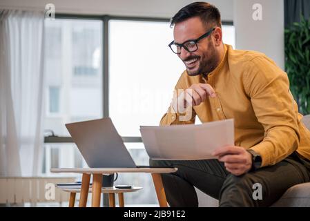Smiling young freelancer reading documents. Businessman analyzing business plan while working on laptop. He is sitting on sofa at home office. Stock Photo