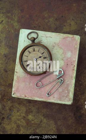 Antique pocket watch lying on scuffed pink cardboard box with two safety pins Stock Photo