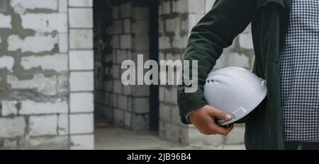 Construction Worker at Site holding helmet close-up. Smart civil architect engineer inspecting and working outdoors building side Stock Photo